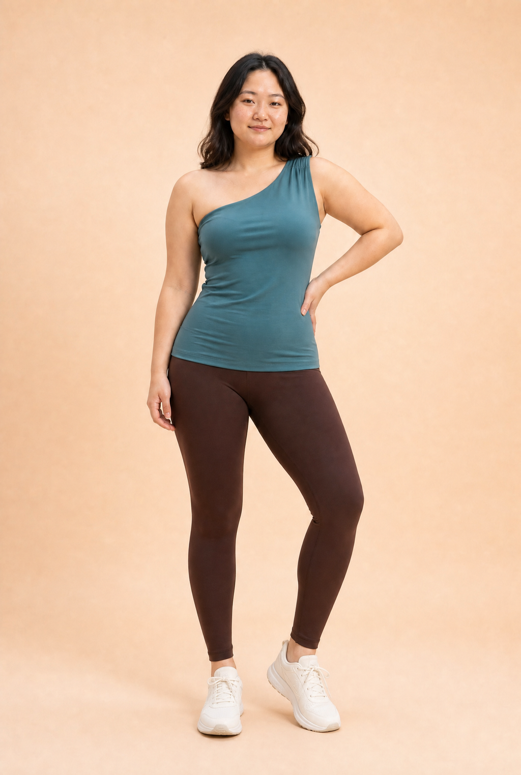 Woman wearing a teal one-shoulder top and brown leggings on a beige background