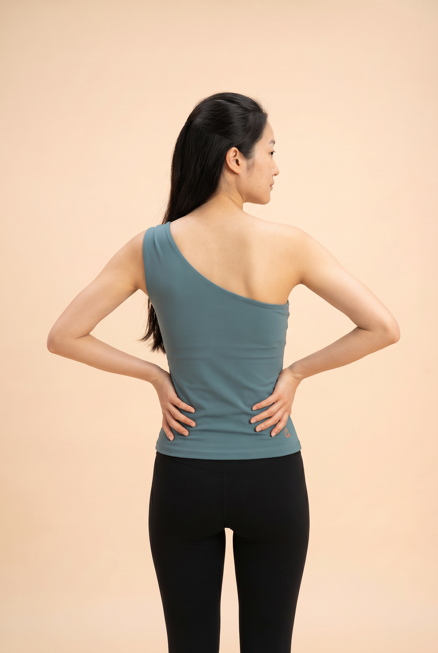 Woman wearing a blue one-shoulder top, showing the garment back view, and black leggings against a beige background